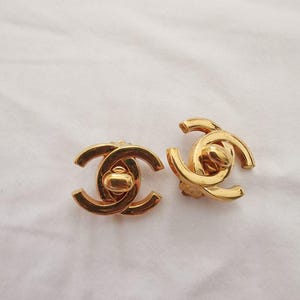 Puede incluir: Un par de pendientes de logotipo de Chanel de color dorado. Los pendientes est&aacute;n hechos de metal y tienen un dise&ntilde;o cl&aacute;sico de CC entrelazado.