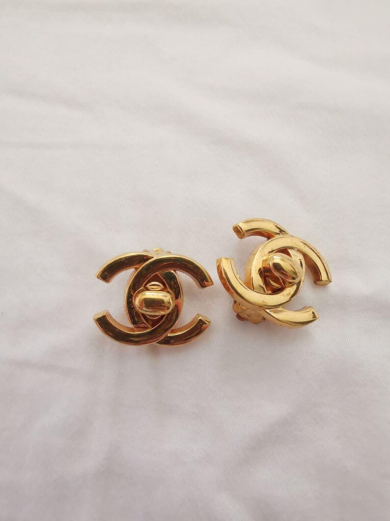 Puede incluir: Un par de pendientes de logotipo de Chanel de color dorado. Los pendientes est&aacute;n hechos de metal y tienen un dise&ntilde;o cl&aacute;sico de CC entrelazado.