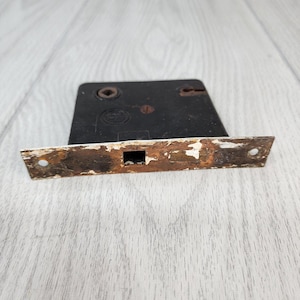 May include: An antique door lock with a rectangular black body and a distressed, rusty metal plate. The plate has a rectangular opening and two screw holes. The lock shows signs of age and wear, with chipped paint and rust.