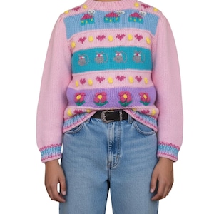May include: A pink knit sweater with a colorful pattern. The sweater features blue, purple, and yellow stripes with embroidered flowers, hearts, and car designs. The sleeves and neckline have a striped trim. The person is wearing blue jeans and a black belt.