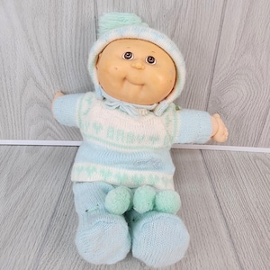 May include: A vintage Cabbage Patch Kid doll wearing a light blue and white knit outfit. The doll has a light skin tone, brown eyes, and a smiling expression. The outfit includes a hat, sweater, and pants with pom-poms.