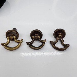 May include: Three antique brass drawer pulls. Each pull features a circular backplate with a decorative, curved handle. The pulls have a warm, aged finish, suggesting vintage or retro design. Ideal for furniture restoration or adding a classic touch.