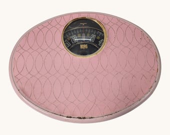 May include: An oval-shaped pink bathroom scale with a repeating dark dotted oval pattern across the surface. The center features a circular analog dial with a gold-toned rim, black background, and the text BORG printed in gold lettering.