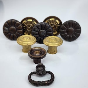 May include: A collection of antique cabinet hardware, including various knobs and a pull. The knobs feature floral and geometric designs in shades of bronze, gold, and dark brown. The pull has an ornate design and a curved handle.