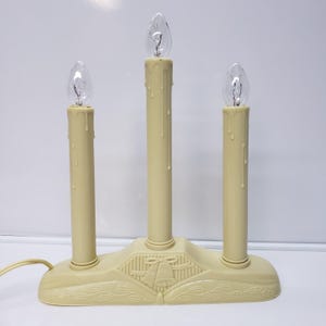 May include: A cream-colored electric candle bridge with three candle-shaped lights. The base features a decorative design with bells and leaves. Each candle has a clear, teardrop-shaped bulb. A power cord extends from the base.