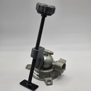 May include: A gray metal bench vise featuring a ball-and-socket joint base, a vertical threaded rod with a black plastic adjustment knob at the top, and a flat black rectangular base plate at the bottom. A small white label reads "nter Tools".