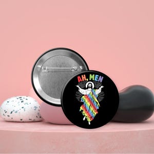 May include: A black button with the text "AH, MEN" and a graphic of a figure in a rainbow robe. The button is displayed with a silver-backed button and other decorative objects on a pink surface.