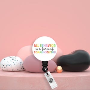 Puede incluir: Un carrete de insignia retráctil blanco con el texto "ALL BEHAVIOR is a form of COMMUNICATION" en colores arcoíris. El carrete es negro y tiene una correa de plástico transparente. El fondo es rosa con piedras decorativas.