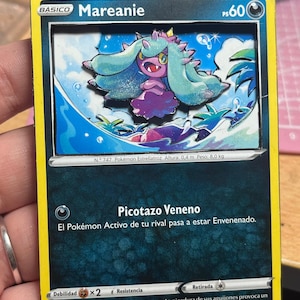 May include: A Mareanie Pokémon card with a 3D effect. The card features a teal and purple creature with the text "Mareanie" and "Picotazo Veneno". The card has a yellow border and includes text in Spanish.