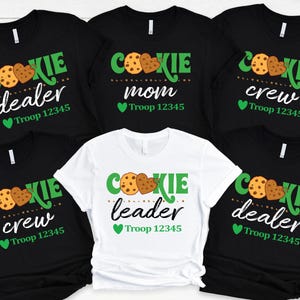 May include: Five black and white t-shirts with green text and images of cookies. The text on the shirts reads "Cookie dealer", "Cookie mom", "Cookie crew", "Cookie crew", and "Cookie leader". Each shirt also includes the text "Troop 12345".
