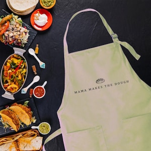 May include: A beige kitchen apron with a pocket and the text "Mama Makes the Dough" in black. The apron has a small illustration of a loaf of bread above the text.