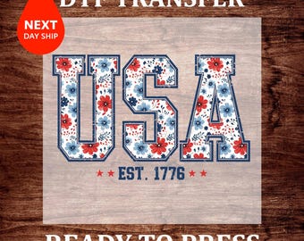 May include: DTF transfer with the word "USA" in a collegiate font, filled with a floral pattern of red and blue flowers. The text "EST. 1776" is below. A red teardrop with "NEXT DAY SHIP" is in the upper left corner.