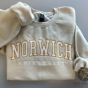 K&ouml;nnte beinhalten: Ein beiger Pullover mit der Aufschrift "Norwich University", die in Gold und Wei&szlig; gestickt ist.