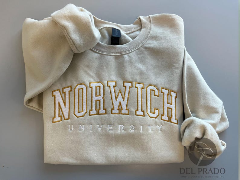 K&ouml;nnte beinhalten: Ein beiger Pullover mit der Aufschrift "Norwich University", die in Gold und Wei&szlig; gestickt ist.