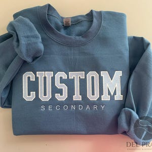 K&ouml;nnte beinhalten: Ein blaues Sweatshirt mit dem Text "CUSTOM SECONDARY" in wei&szlig;en Buchstaben.