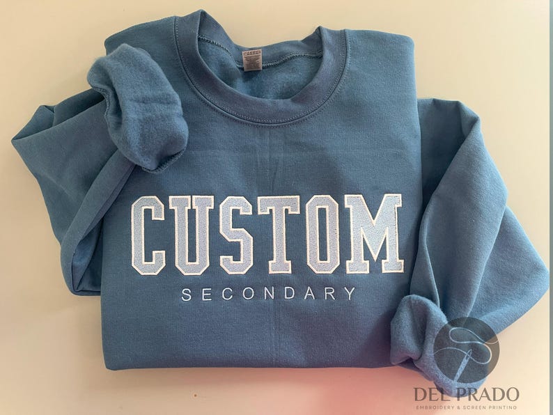 K&ouml;nnte beinhalten: Ein blaues Sweatshirt mit dem Text "CUSTOM SECONDARY" in wei&szlig;en Buchstaben.