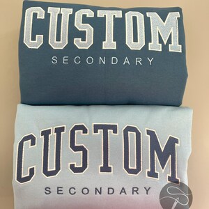 K&ouml;nnte beinhalten: Zwei Sweatshirts mit dem Text "CUSTOM SECONDARY" auf der Vorderseite gestickt. Das obere Sweatshirt ist blaugr&uuml;n und das untere Sweatshirt ist hellblau.