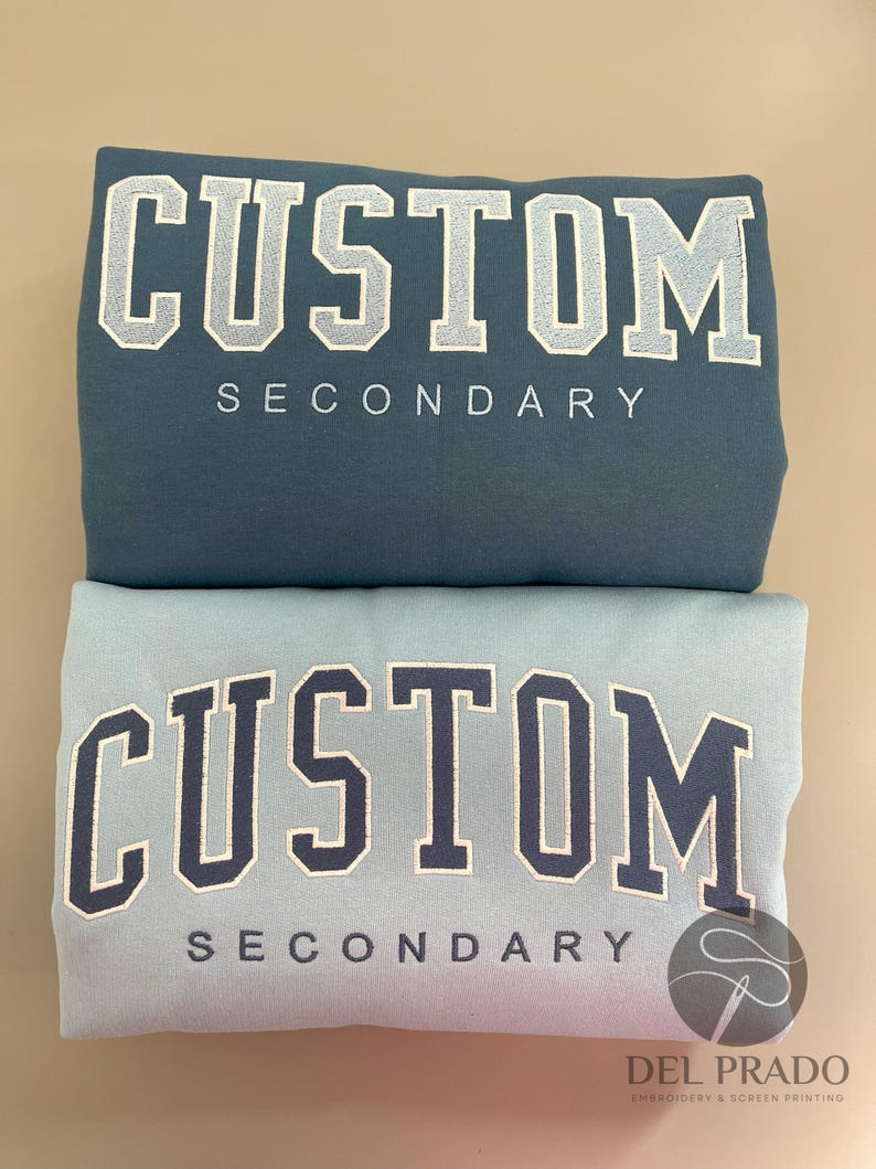 K&ouml;nnte beinhalten: Zwei Sweatshirts mit dem Text "CUSTOM SECONDARY" auf der Vorderseite gestickt. Das obere Sweatshirt ist blaugr&uuml;n und das untere Sweatshirt ist hellblau.