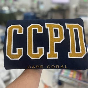 K&ouml;nnte beinhalten: Ein marineblauer Pullover mit den Buchstaben "CCPD" in Gold mit wei&szlig;en Umrandungen. Die Worte "CAPE CORAL" sind in Wei&szlig; unter den Buchstaben gedruckt.