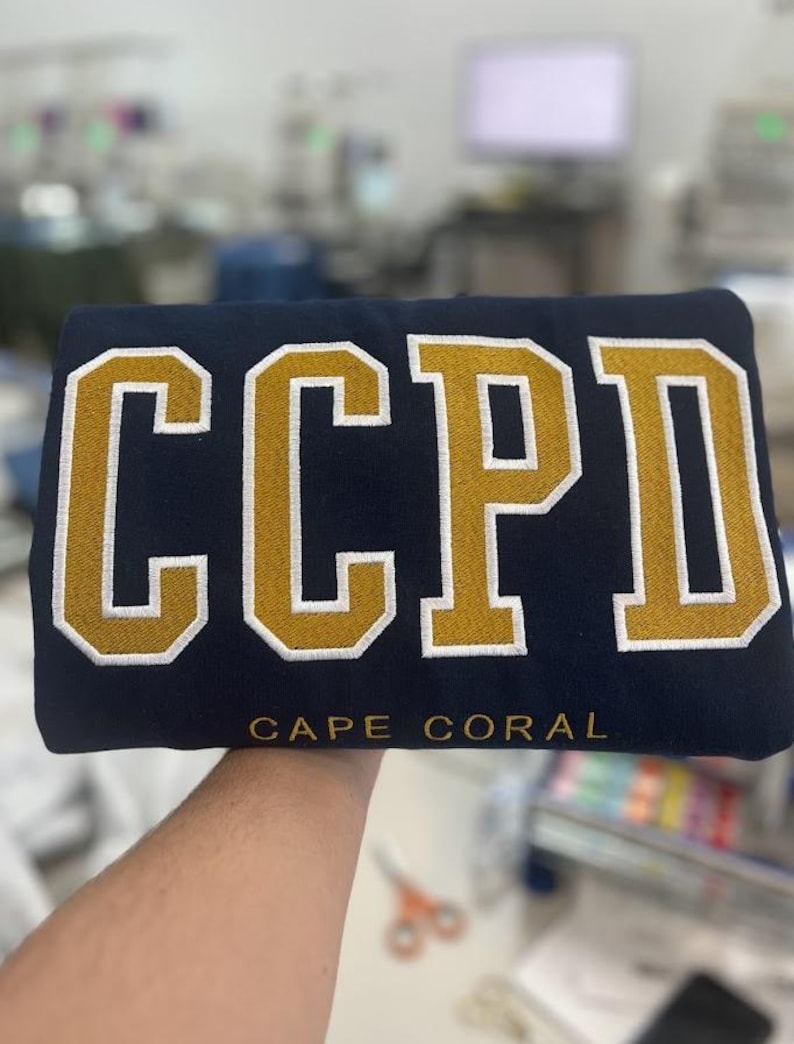 K&ouml;nnte beinhalten: Ein marineblauer Pullover mit den Buchstaben "CCPD" in Gold mit wei&szlig;en Umrandungen. Die Worte "CAPE CORAL" sind in Wei&szlig; unter den Buchstaben gedruckt.