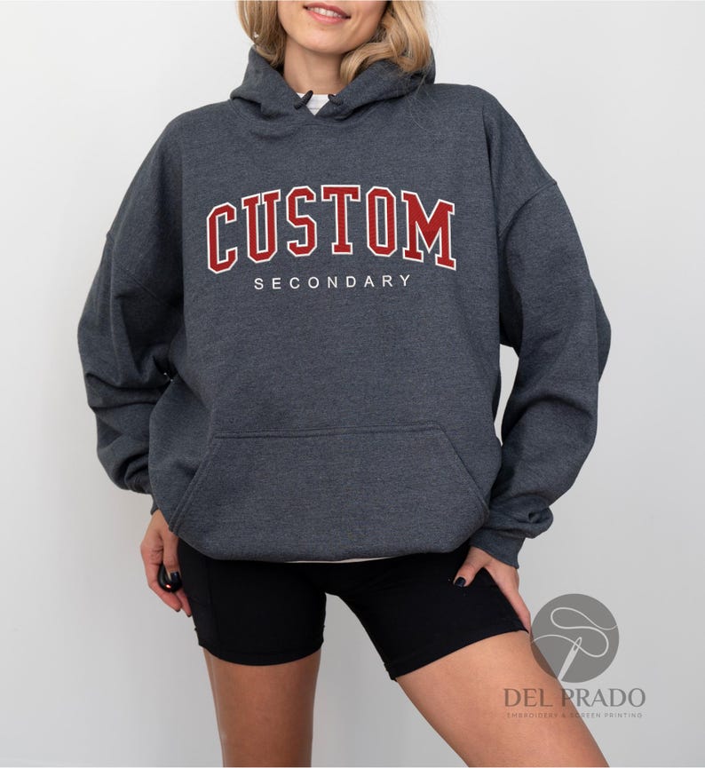 K&ouml;nnte beinhalten: Ein dunkelgrauer Kapuzenpullover mit einem roten "CUSTOM SECONDARY"-Aufdruck auf der Vorderseite.