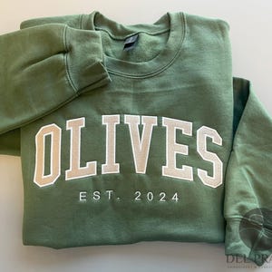 K&ouml;nnte beinhalten: Ein gr&uuml;ner Pullover mit dem Wort "OLIVES" in wei&szlig;en Buchstaben und dem Text "EST. 2024" darunter.