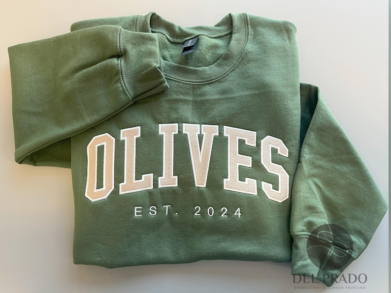 K&ouml;nnte beinhalten: Ein gr&uuml;ner Pullover mit dem Wort "OLIVES" in wei&szlig;en Buchstaben und dem Text "EST. 2024" darunter.