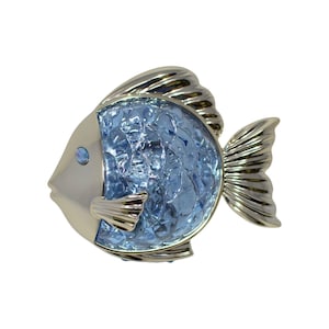 Puede incluir: Broche decorativo con forma de pez, con acabado metálico plateado. El pez presenta un cuerpo facetado azul claro y una pequeña gema azul redonda como ojo. Las aletas y la cola también son plateadas.