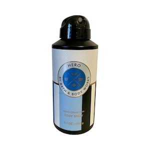 May include: A black and white body spray bottle with a black cap. The label features the word "HERO" in a gold circle with blue accents. The bottle also has the text "MEN'S COLLECTION BODY SPRAY" and is 109 g.
