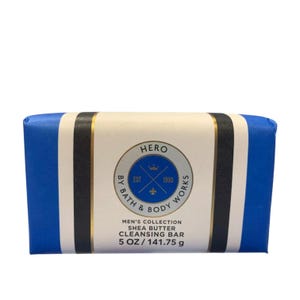 May include: A blue, white, and gold shea butter cleansing bar from the Men's Collection. The bar is wrapped in blue paper with white and gold stripes. The label reads "HERO" and "EST 1990". The bar weighs 5 oz.