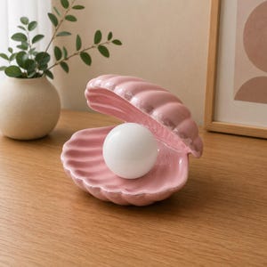 May include: A ceramic lamp shaped like an open seashell in a soft pink colour with a glossy finish. The shell holds a smooth, spherical white light bulb in its centre, mimicking the appearance of a pearl resting inside a clam shell.