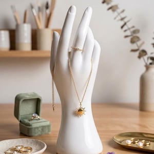 May include: A white ceramic hand-shaped jewelry display stand featuring a gold-toned necklace with a sunburst pendant and a thin gold ring. The display is surrounded by a green velvet ring box, loose pearls, a small purple crystal, and gold rings.