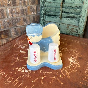 May include: A vintage duck-shaped salt and pepper shaker set. The duck is wearing a blue bonnet and dress with white polka dots. The base is light blue with two white shakers, each with a red letter. A charming kitchen decor item.