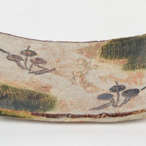 May include: A rectangular ceramic dish with a curved shape. The dish features a beige background with green and brown accents. It is decorated with a floral design in black and brown. The rim is a dark brown color.