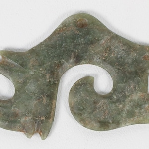 May include: A jade pendant with a unique, abstract design. The green stone has a swirling, open shape with pointed edges and a hole at the top for hanging. The surface shows natural variations in color and texture.