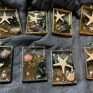 Puede incluir: Diez colgantes rectangulares de resina dorada y plateada con una variedad de conchas y estrellas de mar. Los colgantes están sobre un fondo de tela gris.