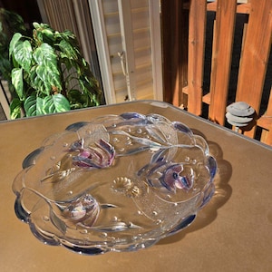 May include: A clear glass serving dish with a scalloped edge and a raised design of flowers and leaves. The dish has a iridescent finish with shades of blue, pink, and gold.