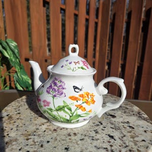 May include: White porcelain teapot with a floral design featuring purple, orange, and pink flowers and butterflies. The teapot has a lid and a handle.