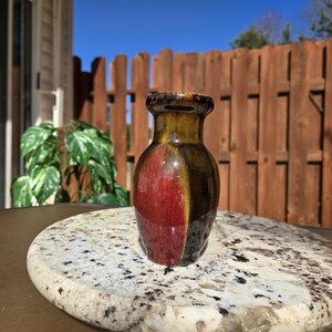 May include: A small, red and brown ceramic vase with a glossy finish. The vase is about 4 inches tall and has a rounded shape.