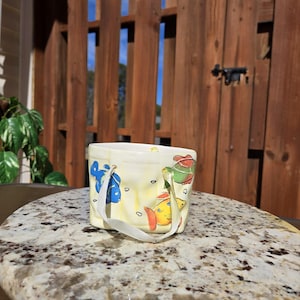 May include: A small white ceramic planter with a handle, featuring a colorful fish design. The planter is sitting on a granite countertop.