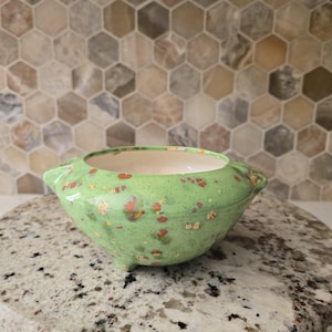 May include: A small, round, green ceramic bowl with three small feet and two handles. The bowl is speckled with orange and yellow spots. The interior of the bowl is a cream color. The bowl is sitting on a speckled gray surface.