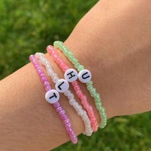 May include: Four colourful beaded bracelets with letter charms spelling out "THUL". The bracelets are stacked on a person's wrist.