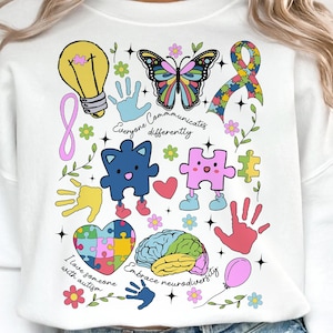 May include: White sweatshirt with colourful autism awareness graphics. Features a lightbulb, butterfly, puzzle pieces, handprints, and the phrases "Everyone Communicates Differently," "I love someone with autism," and "Embrace neurodiversity."