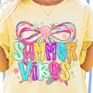 May include: Pale yellow t-shirt featuring the words "Summer Vibes" in a vibrant, multi-coloured design. The graphic includes a bow, seashells, and stars. The text is in bright colours, including pink, blue, and orange.