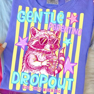 Puede incluir: Una camiseta lila Comfort Colors con un diseño de mapache con gafas de sol y una bebida. La camiseta tiene las palabras "GENTLE PARENTING DROPOUT" y "EST. THIS MORNING" en una fuente divertida.