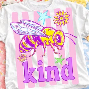 Pode incluir: Camiseta branca com um gráfico colorido de uma abelha, flores e estrelas, sobre listras verticais rosa. A palavra "kind" é impressa em roxo na parte inferior. A camiseta é exibida em um fundo com estampa floral.