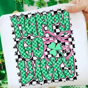 Könnte beinhalten: Weißes T-Shirt zum St. Patrick's Day mit den Worten "Happy Go Lucky" in grün karierten Buchstaben mit rosa Akzenten. Eine rosa Schleife und Kleeblätter sind ebenfalls im Design enthalten. Das T-Shirt wird von einer Hand gehalten.