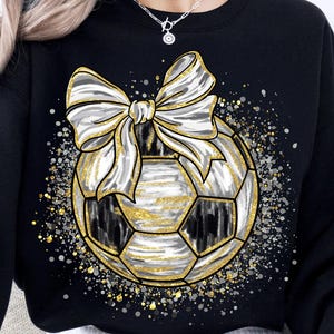 K&ouml;nnte beinhalten: Schwarzes Sweatshirt mit einem Fu&szlig;ball-Design und einer gro&szlig;en Schleife. Der Fu&szlig;ball ist schwarz und wei&szlig; mit goldenem Glitzer. Die Schleife ist wei&szlig; und gold, mit goldenem Glitzer und silbernen Sprenkeln um den Ball.