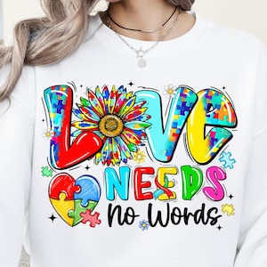 May include: White sweatshirt with the words "LOVE NEEDS no WORDS" in colorful puzzle piece letters. A sunflower and a heart made of puzzle pieces are also featured in the design.