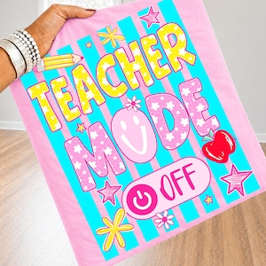 Op de afbeelding: Een roze deken met blauwe verticale strepen en de tekst "TEACHER MODE OFF" in een speels lettertype. Het ontwerp bevat een lachend gezicht, sterren, bloemen, een potlood en een appel. De deken wordt door een persoon vastgehouden.